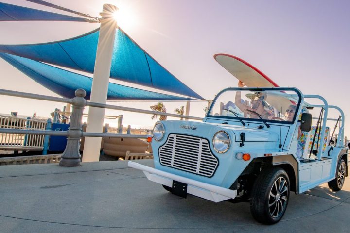Light blue open-top car with surfboards at a sunny waterfront.