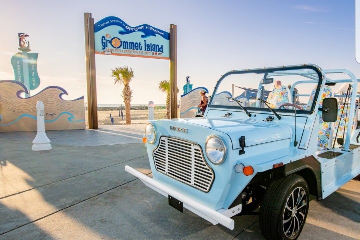 Blue vintage car parked near a beach entrance sign under a clear sky.