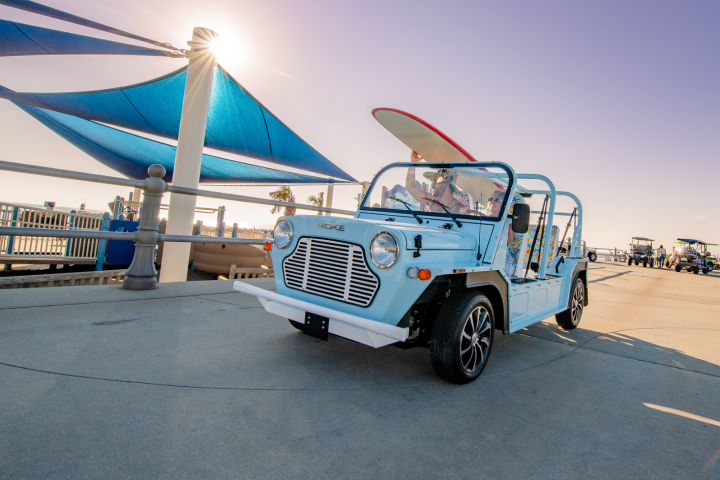 Blue open-air car with surfboard on a sunny pier.