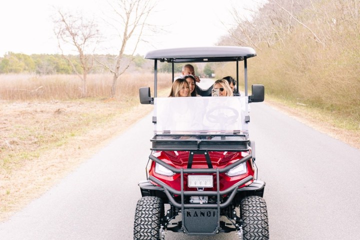 Four people in a golf cart driving on a rural road.