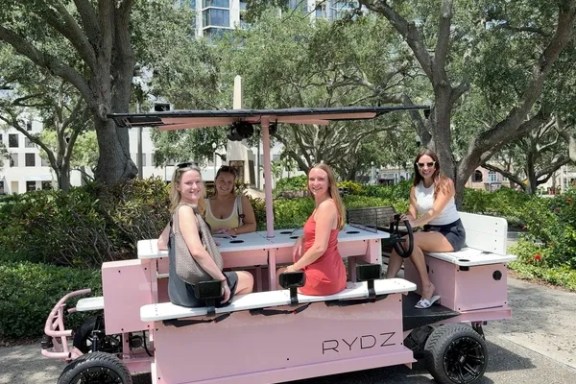 Four women sitting on a pink pedal-powered vehicle named 'RYDZ' outdoors.