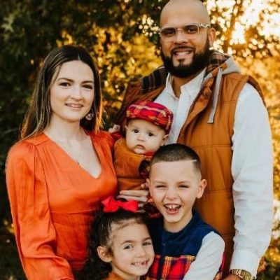Family of five smiling in fall attire with trees in the background.