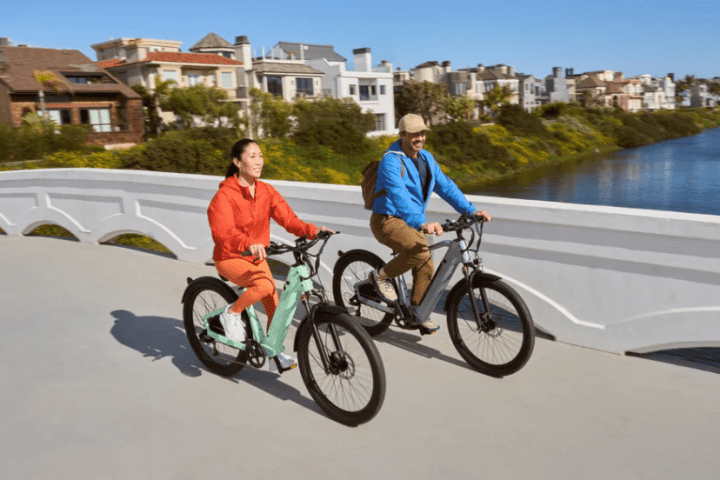 Two people riding bikes on a bridge over a canal, with houses and greenery in the background.