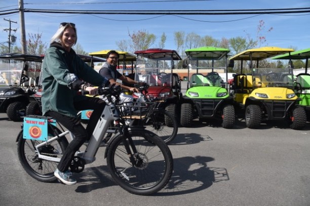 Two people bike past parked colorful golf carts on a sunny day.