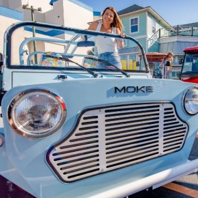 Woman standing on blue Moke car parked in sunny street.