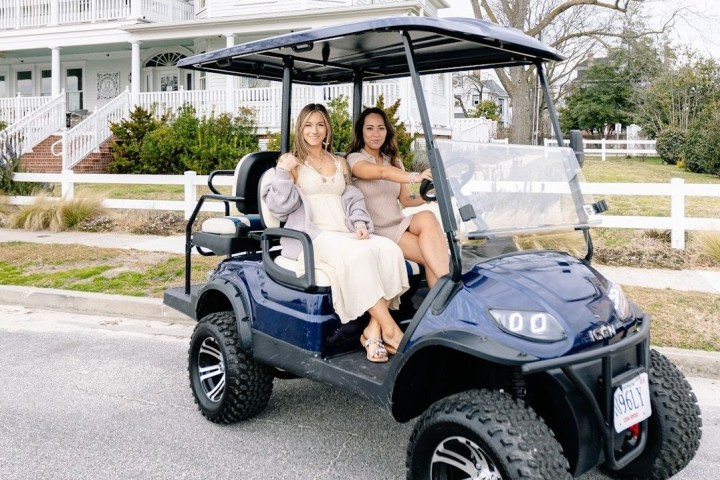 Two women in dresses sit on a blue golf cart by a white fence.